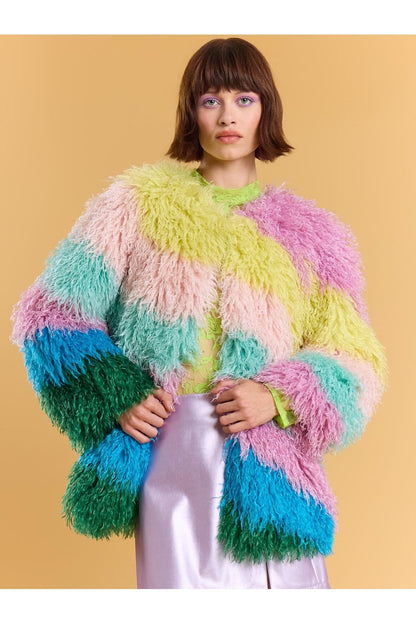 Woman wears a pastel rainbow faux fur coat with bright stripes over a shiny lavender skirt, standing against a plain beige background.