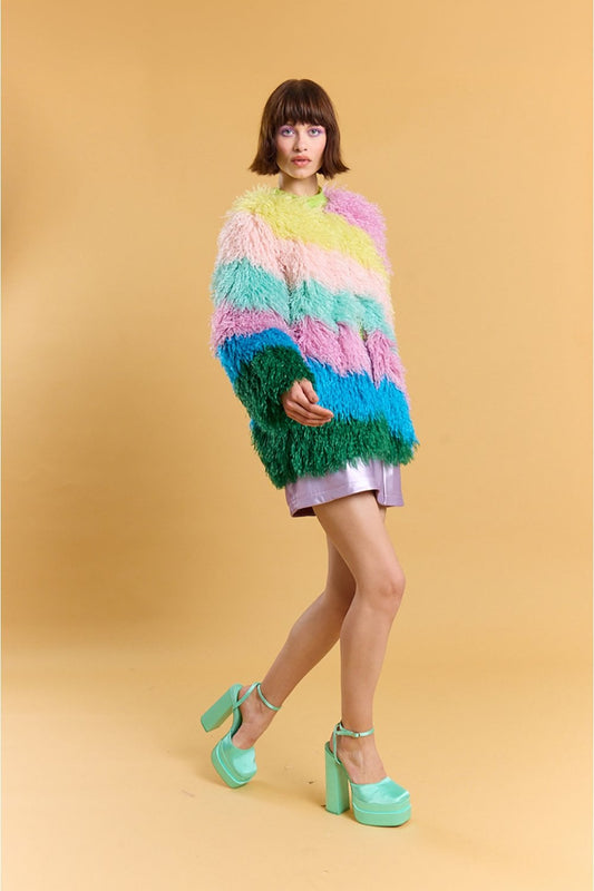 A person stands against a beige background wearing a pastel rainbow faux fur coat, metallic pink shorts, and mint green platform heels.
