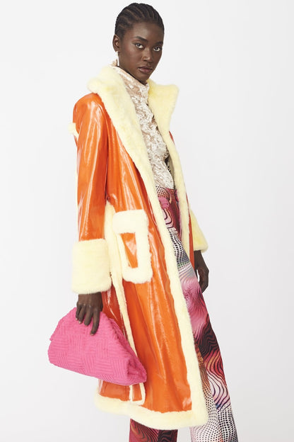A person wearing an orange faux suede trench coat with yellow faux fur trim over a patterned outfit holds a bright pink textured clutch bag, standing against a plain white background.