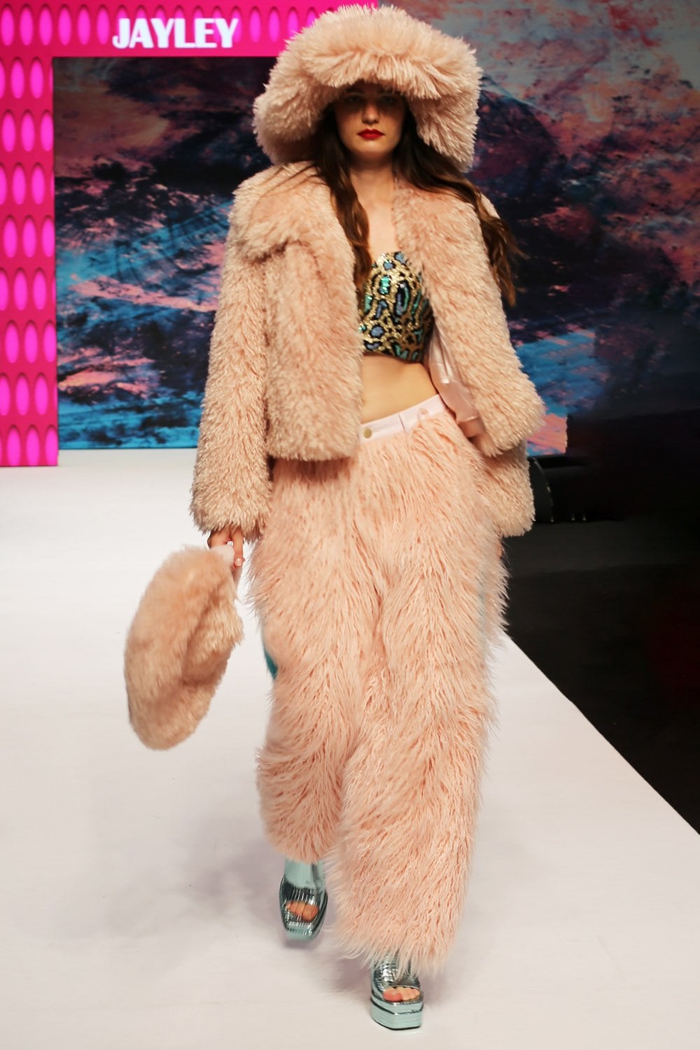 A model walks the catwalk in a pink faux fur jacket and trousers, a patterned crop top, platform sandals, and a luxury faux Mongolian fur hat, carrying a matching pink furry bag.