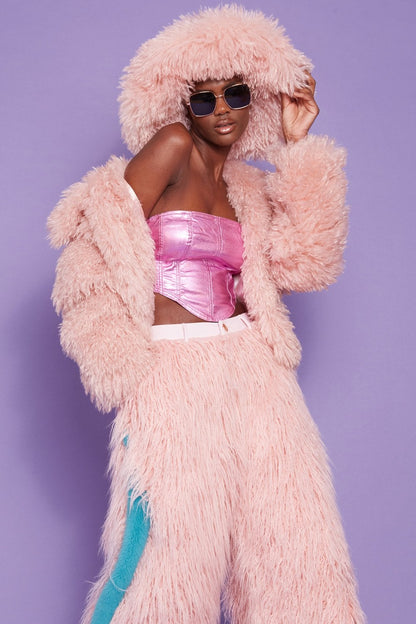 Person wearing a fluffy pink jacket with a hood, pink furry trousers featuring a blue stripe, metallic pink top, sunglasses and an oversized fur hat, standing against a purple background.