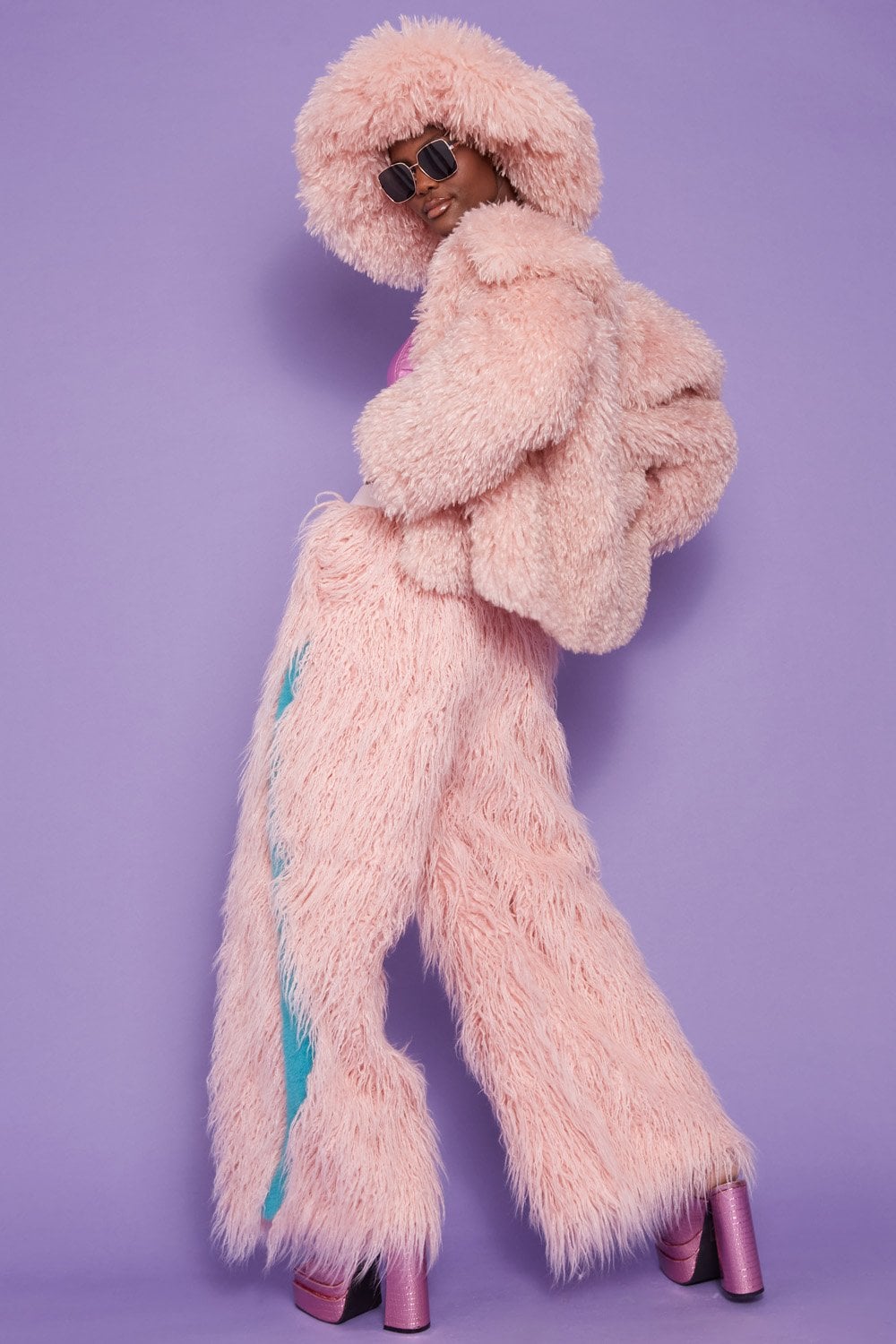 Person in a pink fluffy jacket, matching trousers, luxury faux Mongolian fur hat, hood, and sunglasses poses against a purple background wearing platform shoes.
