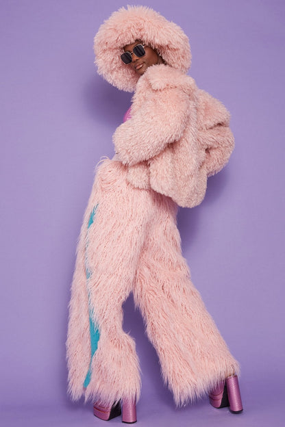 Person in a pink fluffy jacket, matching trousers, luxury faux Mongolian fur hat, hood, and sunglasses poses against a purple background wearing platform shoes.
