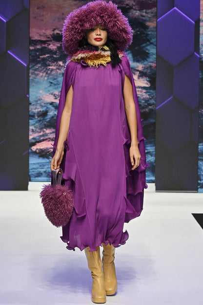 Model on catwalk wearing a loose purple dress, vegan faux fur hat and bag, beige boots, and a layered necklace, set against a geometric and abstract backdrop.