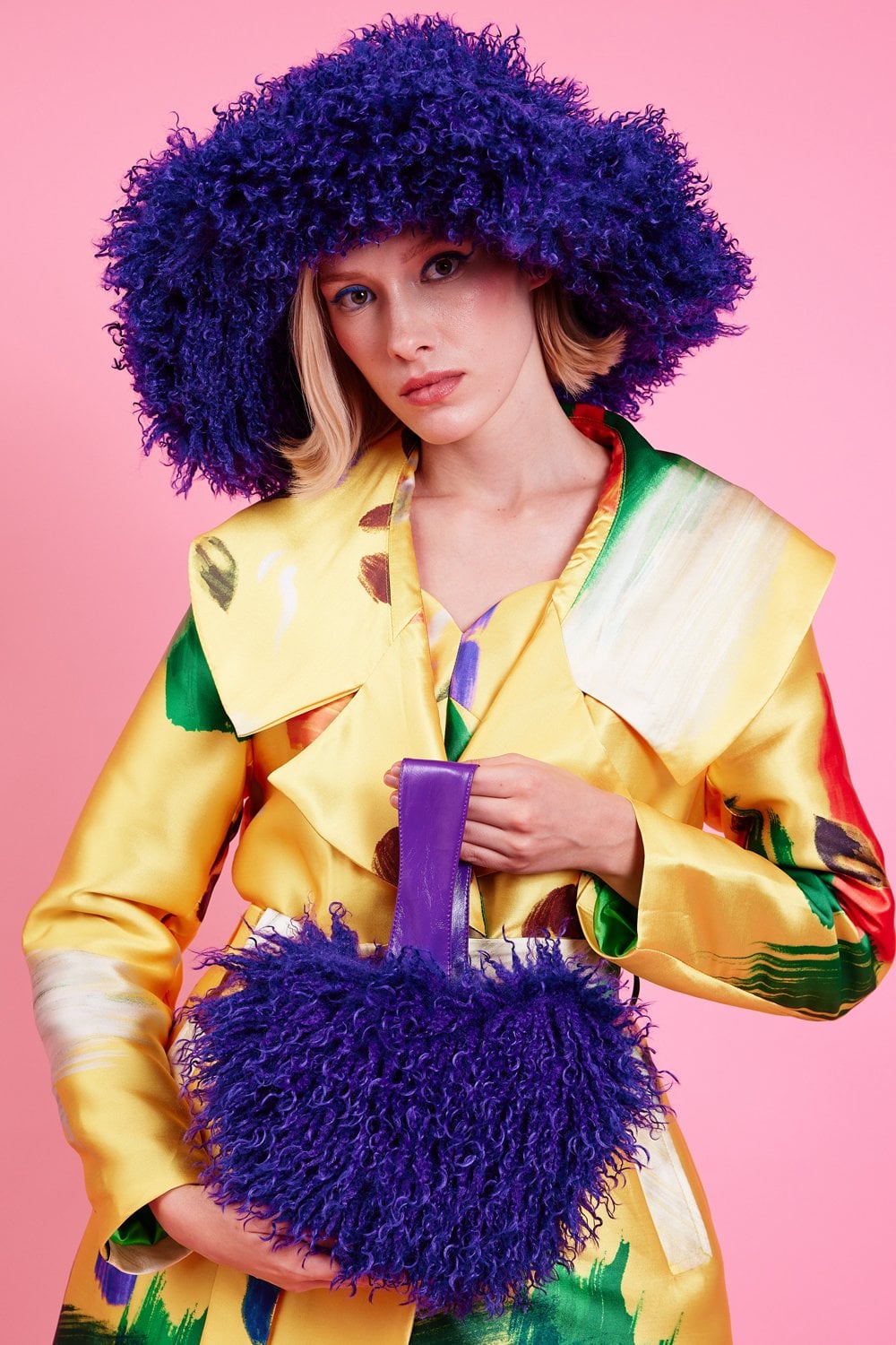 A woman in a yellow patterned coat holds a matching purple fluffy handbag and wears an oversized vegan faux fur hat against a pink background.