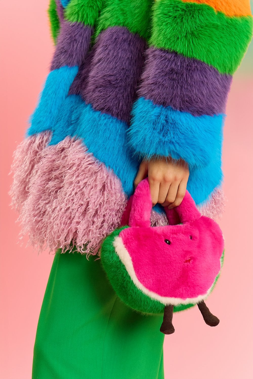 A person in a colourful faux fur coat and green trousers holds a playful faux fur bag shaped like a watermelon slice, set against a vibrant pink background.