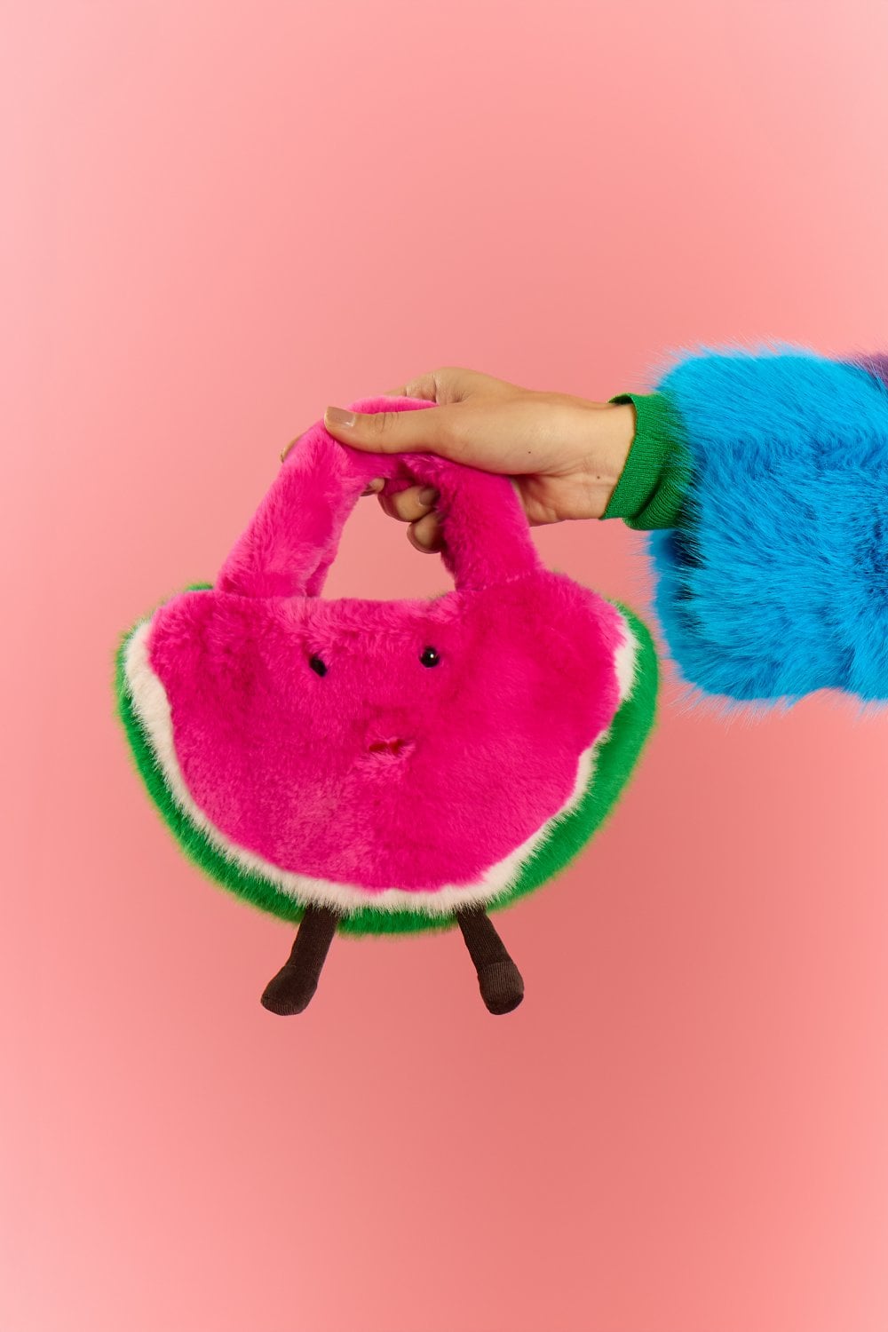 A person wearing a colourful faux fur sleeve holds a plush watermelon bamboo bag shaped like a pink slice with a face and legs, set against a pink background.