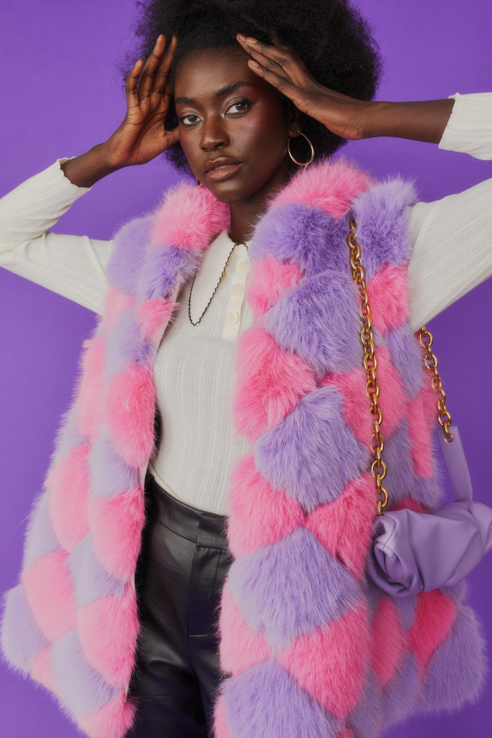 A person wearing a pink and purple chequered faux fur gilet, white long-sleeved shirt, and black trousers stands against a purple background holding a lavender handbag with a chain strap.