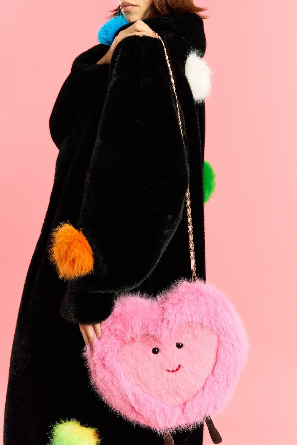 A person in a black coat adorned with colourful pom-poms holds a pink faux fur heart bag featuring a smiling face, set against a solid pink background.