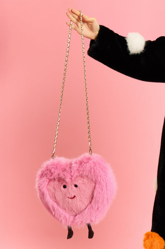 Someone holds a faux fur heart bag with a smiling face, gold chain strap, and small legs, set against a pink background.