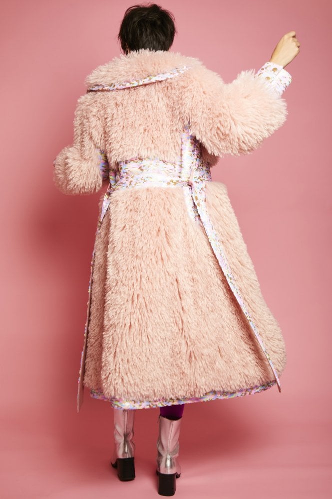 Person seen from behind, wearing a long pink faux Mongolian fur coat with a belted waist and metallic boots, standing against a pink backdrop.