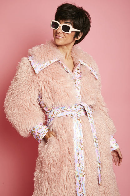 Person in a fluffy pink faux Mongolian fur coat with sequin belt and cuffs, paired with white square sunglasses, poses against a pink backdrop.