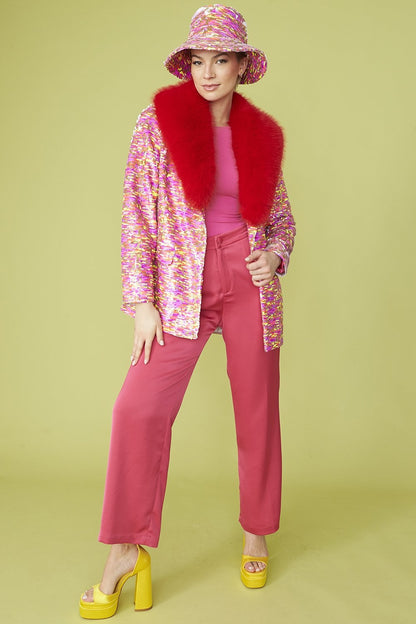 Woman posing in a colourful outfit with a glamorous blazer featuring a red faux fur collar, matching hat, pink top and trousers, and yellow platform heels against a yellow background.