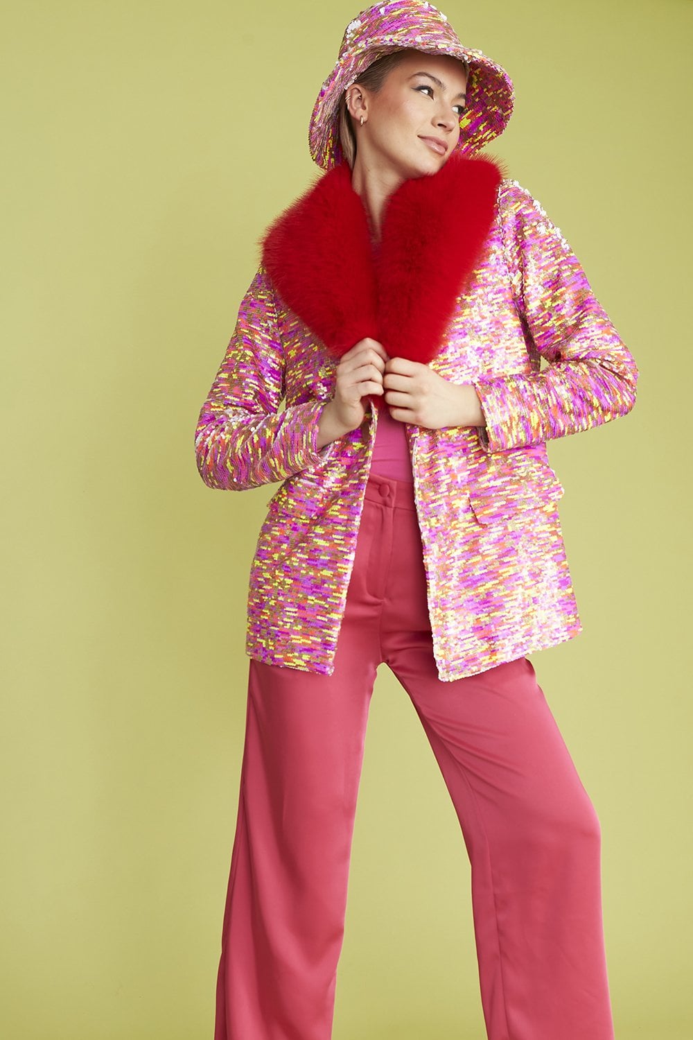 Person wearing a colourful textured jacket with a red faux fur collar, matching wide-leg pink trousers, and a bucket hat, standing against a light green background.