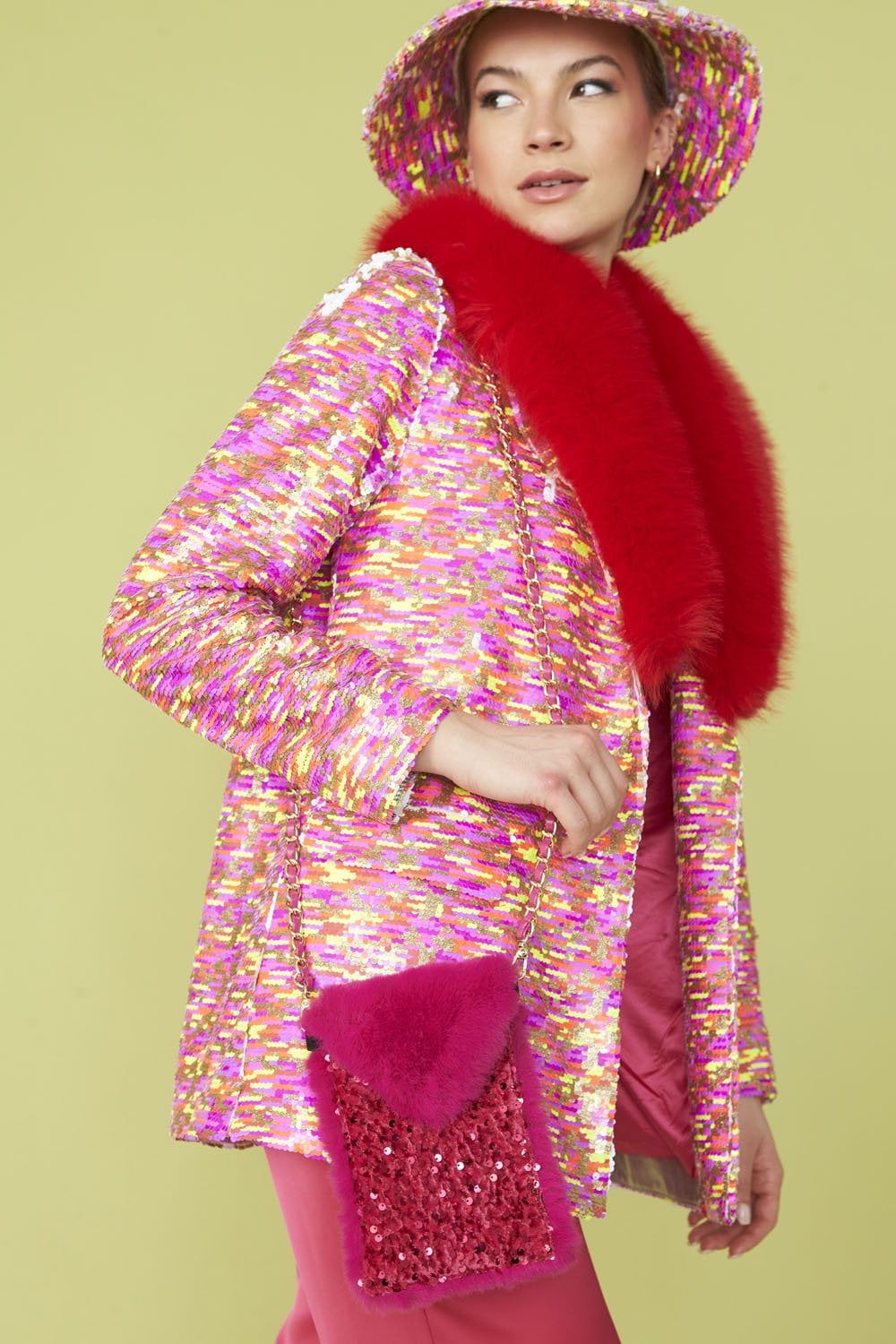 A woman wears a glamorous blazer with a red faux fur collar, matching hat and pink trousers, holding a pink and red sequinned handbag against a light green background.