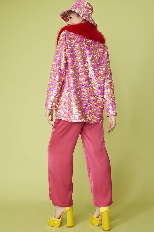 A person stands with their back to the viewer, dressed in a glamorous blazer featuring a red faux fur collar, pink trousers, yellow platform heels, and a matching hat, set against a pale green background.