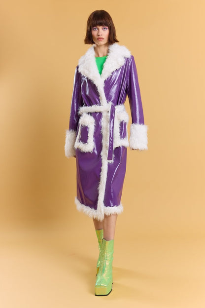 A person stands against a tan background wearing a shiny royal purple faux leather coat with white faux fur trim, a green top, and metallic green platform boots.