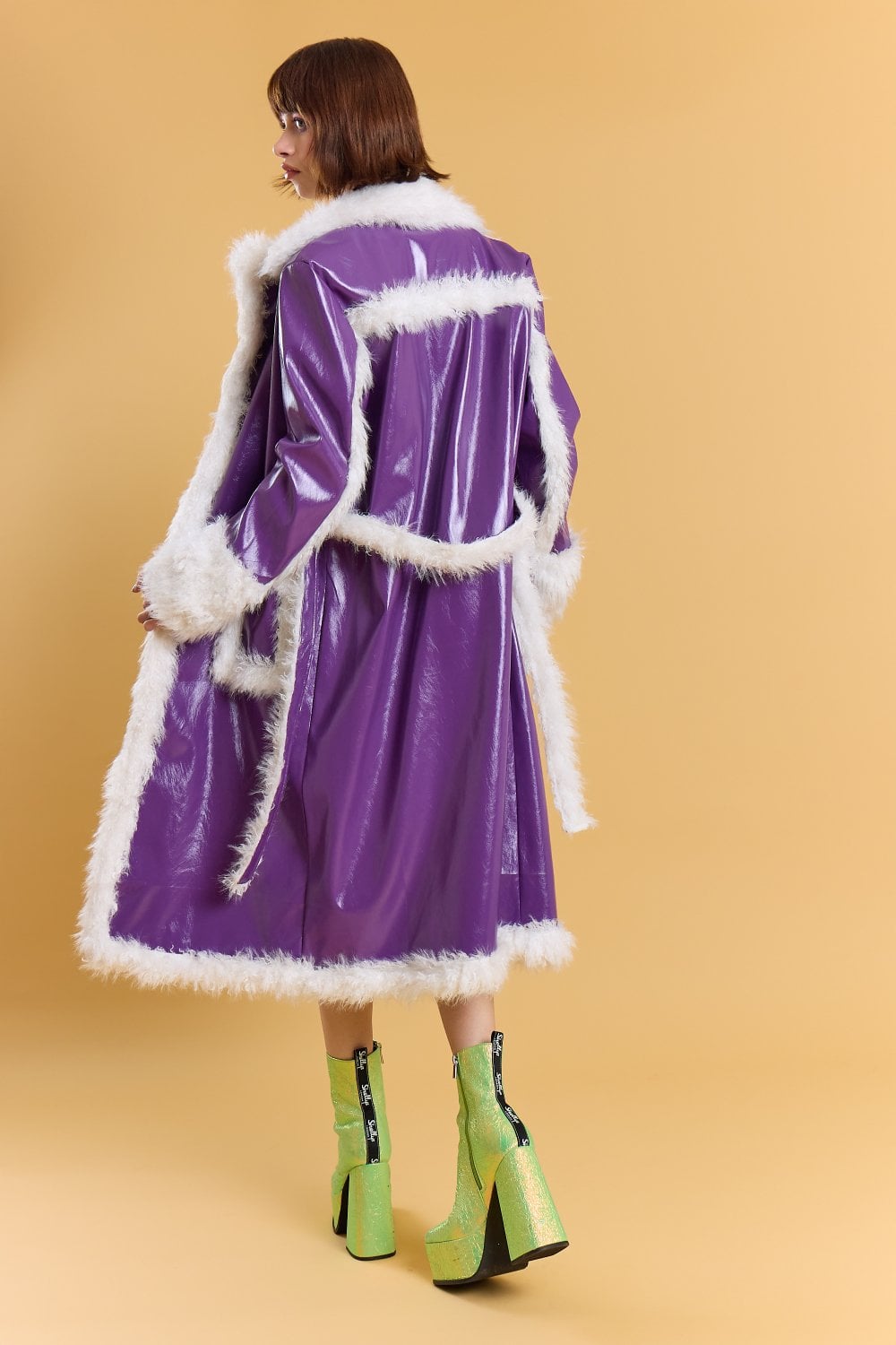 Person in a long, royal purple trench coat with white faux fur trim and bright green high platform boots, standing against a plain beige background.