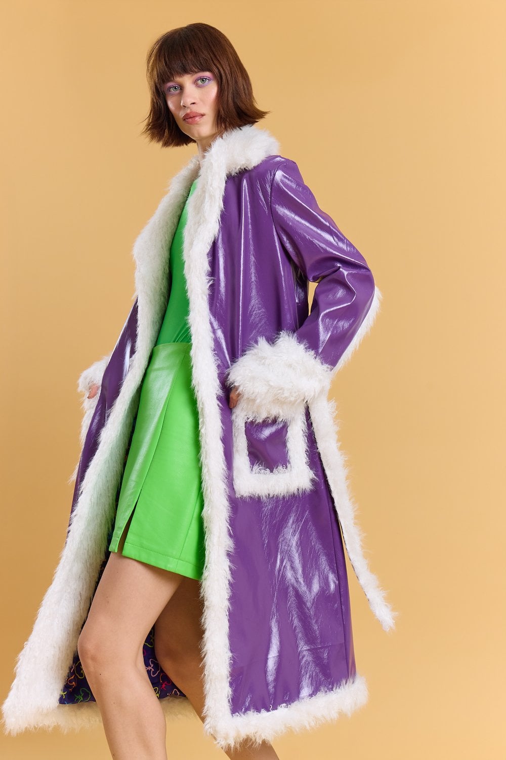 A person in a shiny purple faux leather trench coat with white faux fur trim over a vibrant green dress stands against a plain beige backdrop.