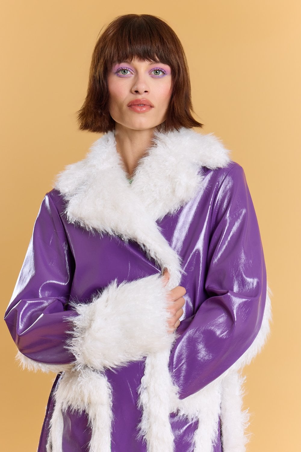 A person with short brown hair wears a shiny purple faux leather coat with white faux fur trim, standing against a plain beige background.