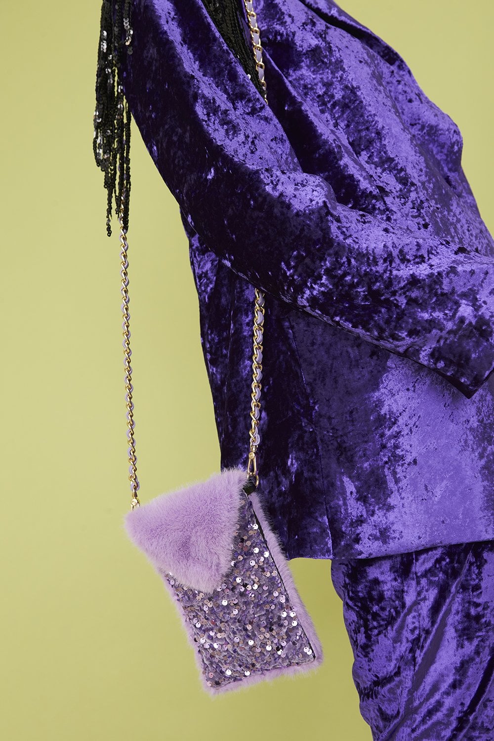 Person wearing a purple crushed velvet outfit with a lavender faux fur and sequin mobile holder bag on a gold chain, set against a pale green background.
