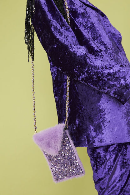 Person wearing a purple crushed velvet outfit with a lavender faux fur and sequin mobile holder bag on a gold chain, set against a pale green background.