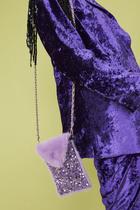 Person wearing a purple crushed velvet outfit with a lavender faux fur and sequin mobile holder bag on a gold chain, set against a pale green background.