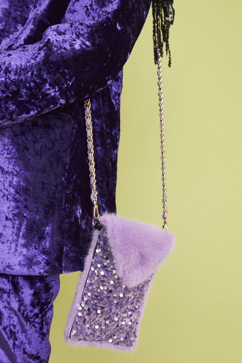 A person in a purple velvet outfit holds a small, stylish mobile bag crafted from lavender faux fur and sequins, featuring a gold chain strap, set against a light green background.