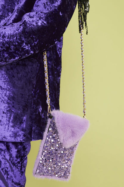 A person in a purple velvet outfit holds a small, stylish mobile bag crafted from lavender faux fur and sequins, featuring a gold chain strap, set against a light green background.