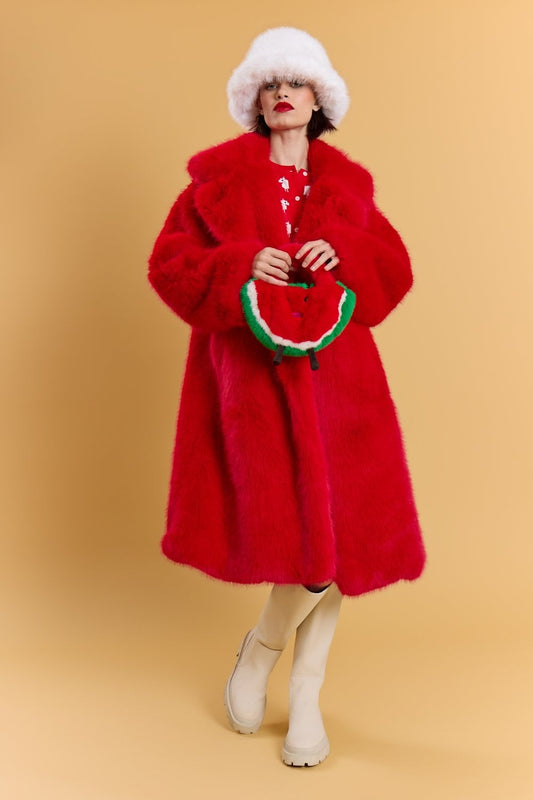 A person dressed in a bright red fur coat, a white fluffy hat, white boots, and holding a watermelon-shaped bamboo bag stands against a beige background.