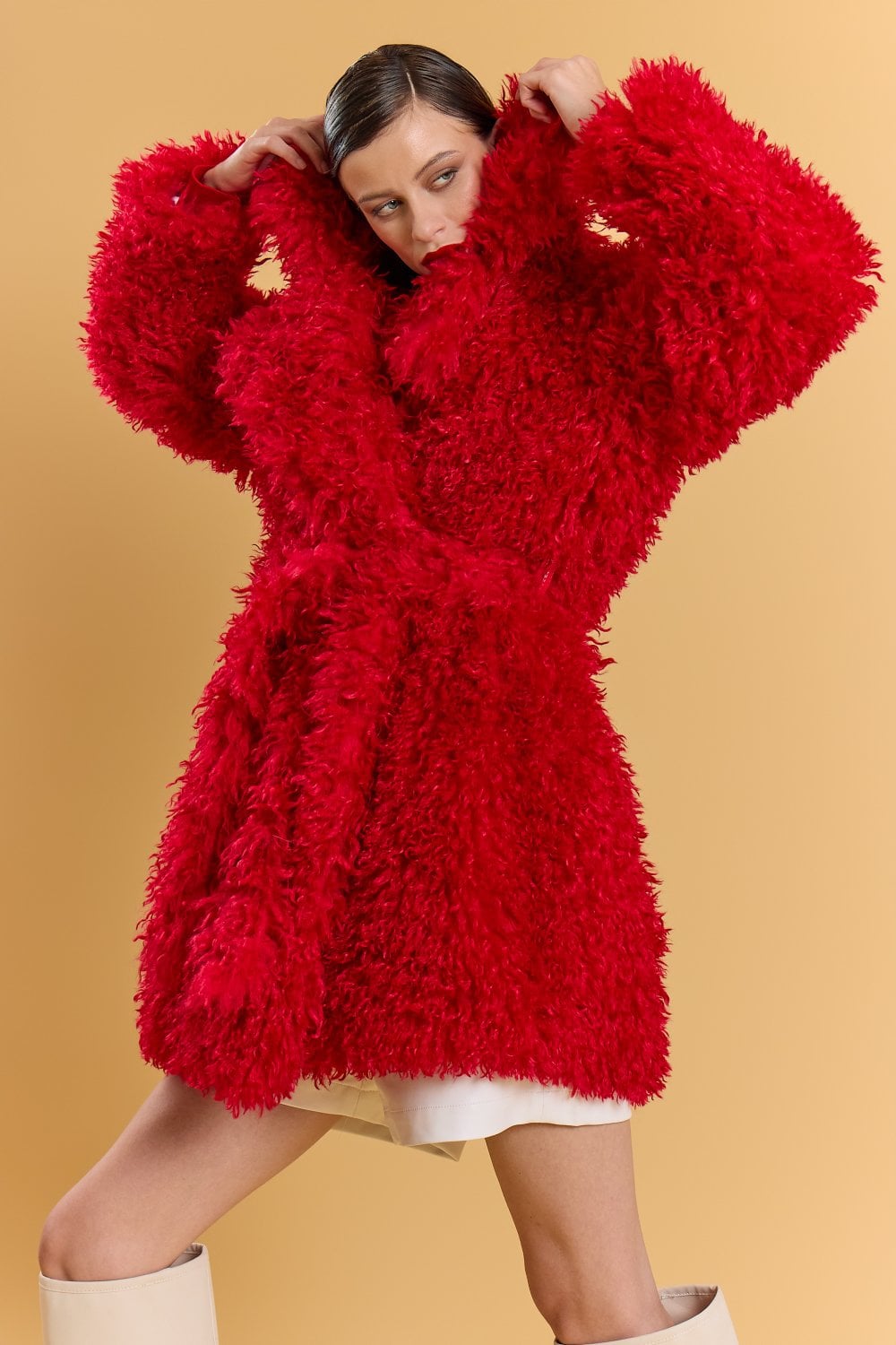 Woman poses in a vibrant red faux fur coat with a belt, white mini skirt, and knee-high white boots against a beige backdrop. The red faux fur coat brings striking texture and warmth to her stylish ensemble.