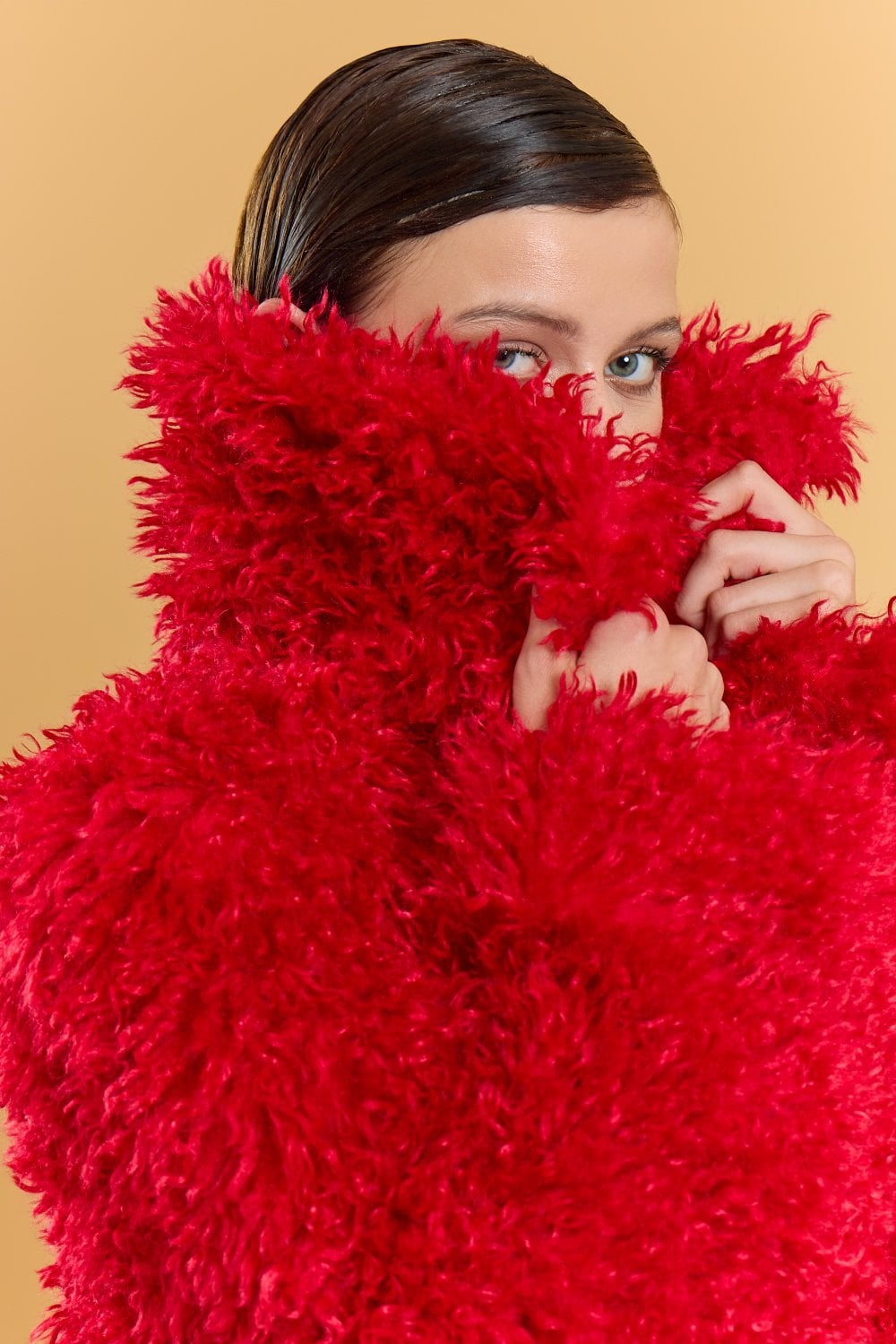 A person wearing a bright red bamboo blend faux fur coat pulls the collar over their lower face, with only their eyes visible; the background is a plain beige colour.