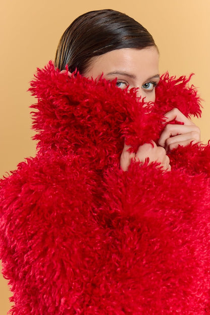 A person wearing a bright red bamboo blend faux fur coat pulls the collar over their lower face, with only their eyes visible; the background is a plain beige colour.