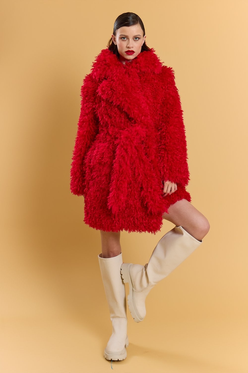 A woman poses against a beige background, wearing a striking red faux fur coat with bold texture and knee-high white boots.