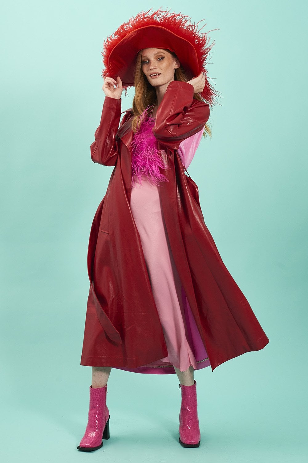 A woman poses in a red eco leather trench coat, pink dress, and pink boots, wearing a large red hat with feather trim against a plain light blue background.