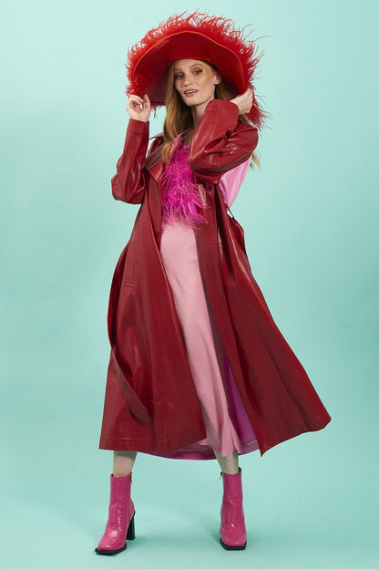 A woman poses in a red eco leather trench coat, pink dress, and pink boots, wearing a large red hat with feather trim against a plain light blue background.