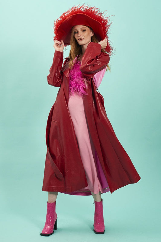 A woman poses in a red eco leather trench coat, pink dress, and pink boots, wearing a large red hat with feather trim against a plain light blue background.
