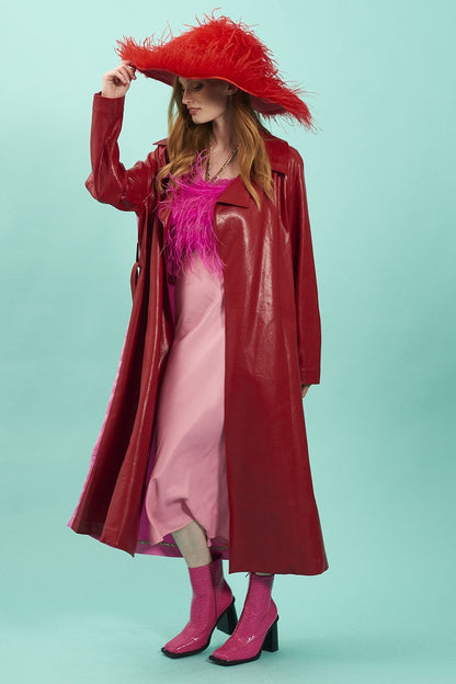 A person wearing a red eco leather trench coat, large red feathered hat, pink dress, and pink ankle boots stands against a light turquoise background.