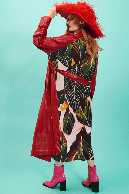 A woman stands against a turquoise background, wearing a red eco leather trench coat adorned with feathers, a tropical print dress, and pink boots.