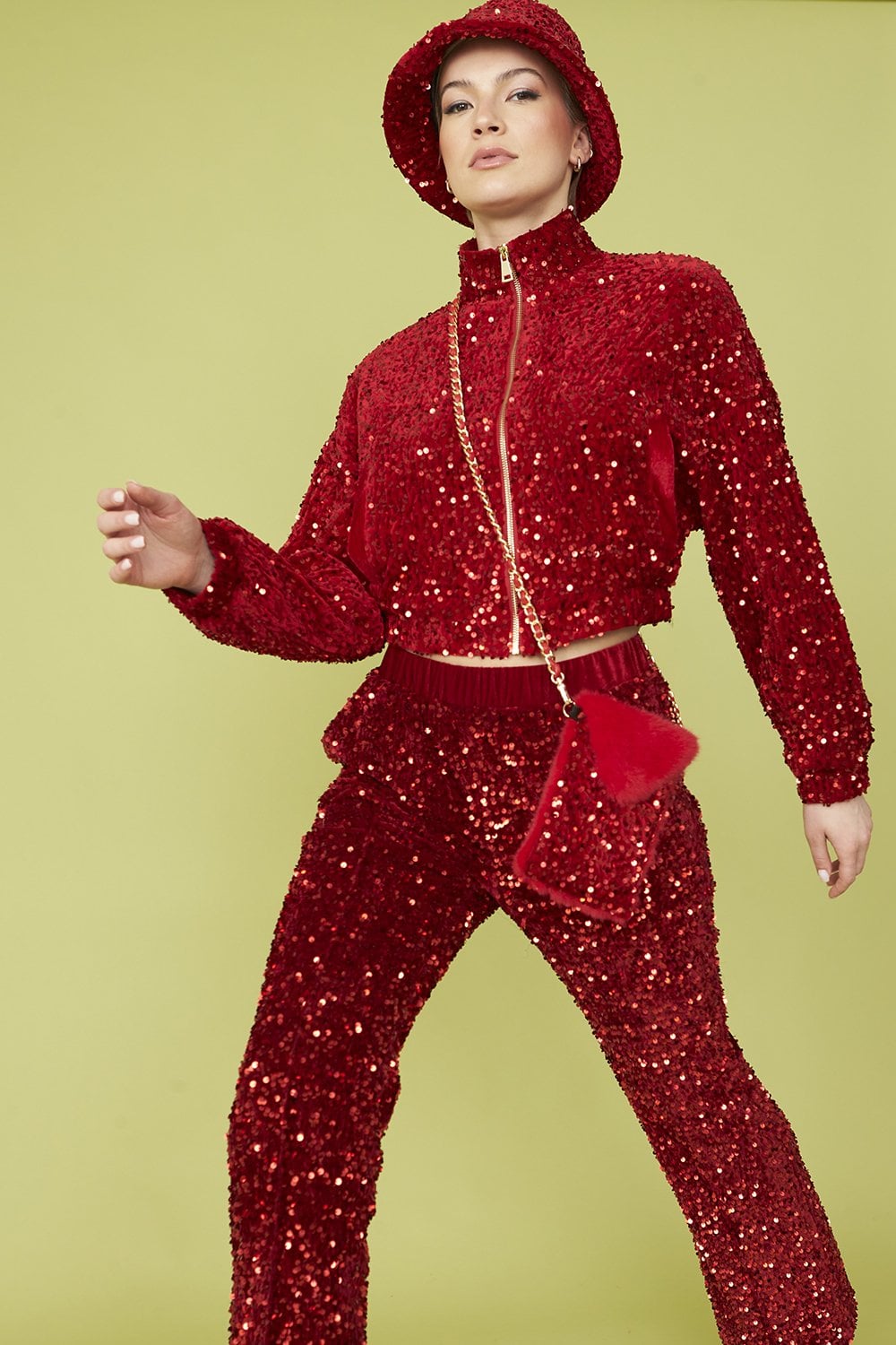 Person dressed in a red sequinned outfit with matching trousers, jacket, bucket hat, and a stylish Sequin Phone Holder Bag, standing against a pale green background.