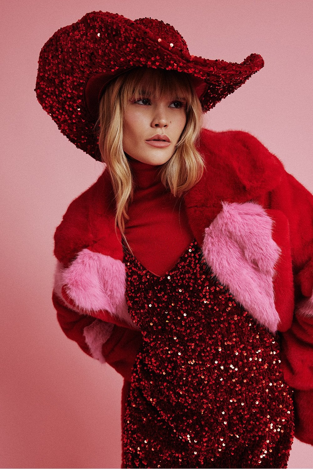 A person dressed in a red sequinned outfit, a red faux fur jacket with pink accents, and a large handmade flapper hat with silk lining poses against a pink background.