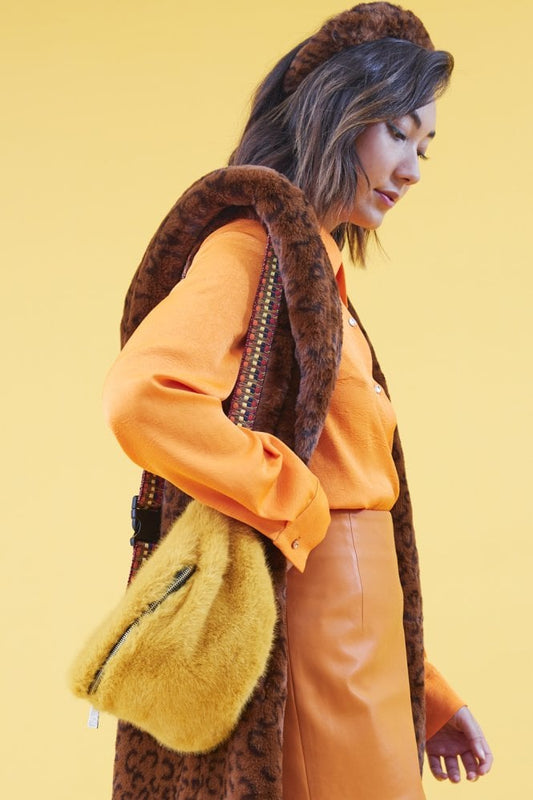 A woman stands against a yellow background, dressed in an orange shirt, brown patterned waistcoat, orange skirt and headband, carrying a large yellow faux fur festival bag over her shoulder.