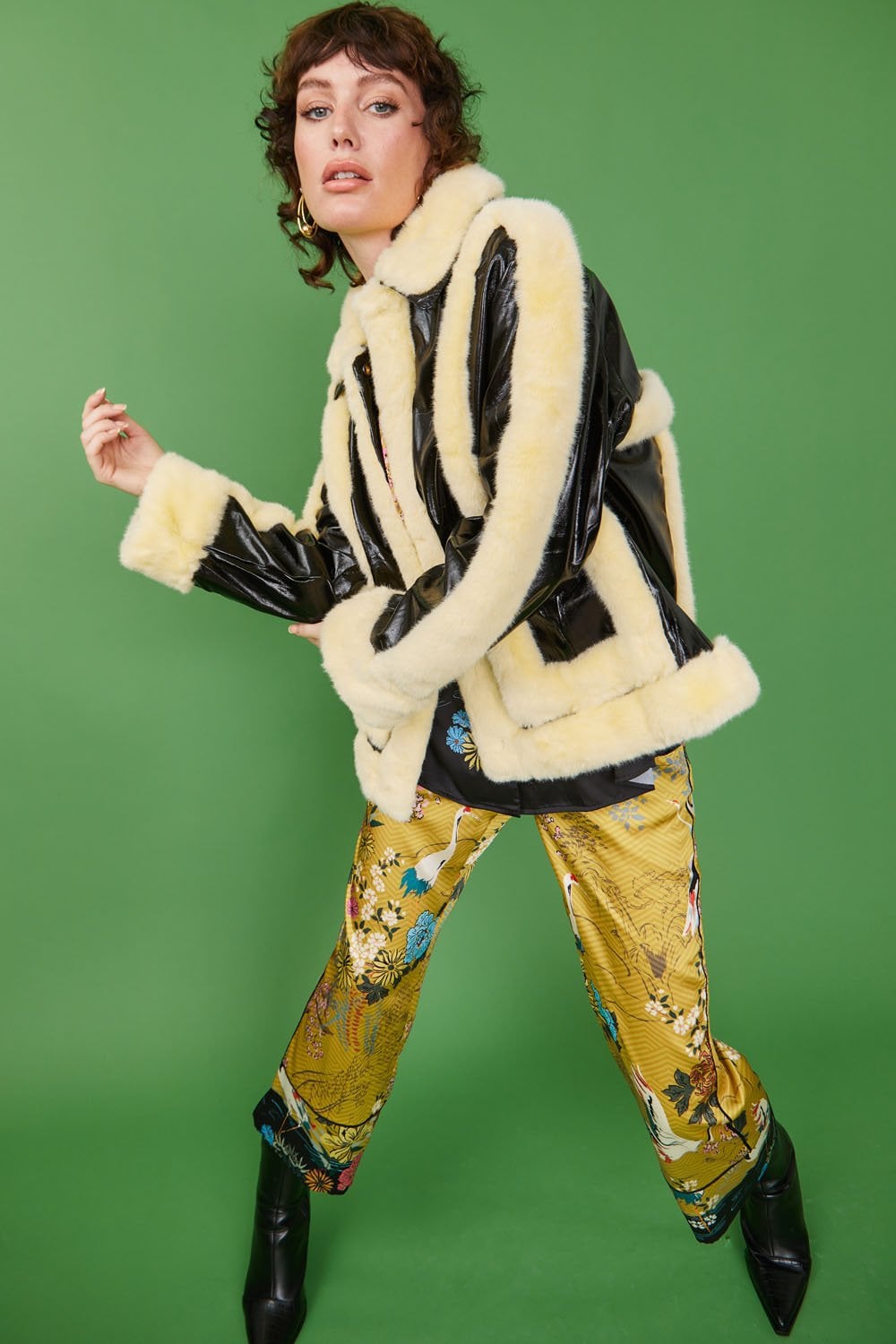 Person wearing a black and cream retro aviator jacket with a shearling collar, patterned yellow trousers, and black boots poses against a green background.