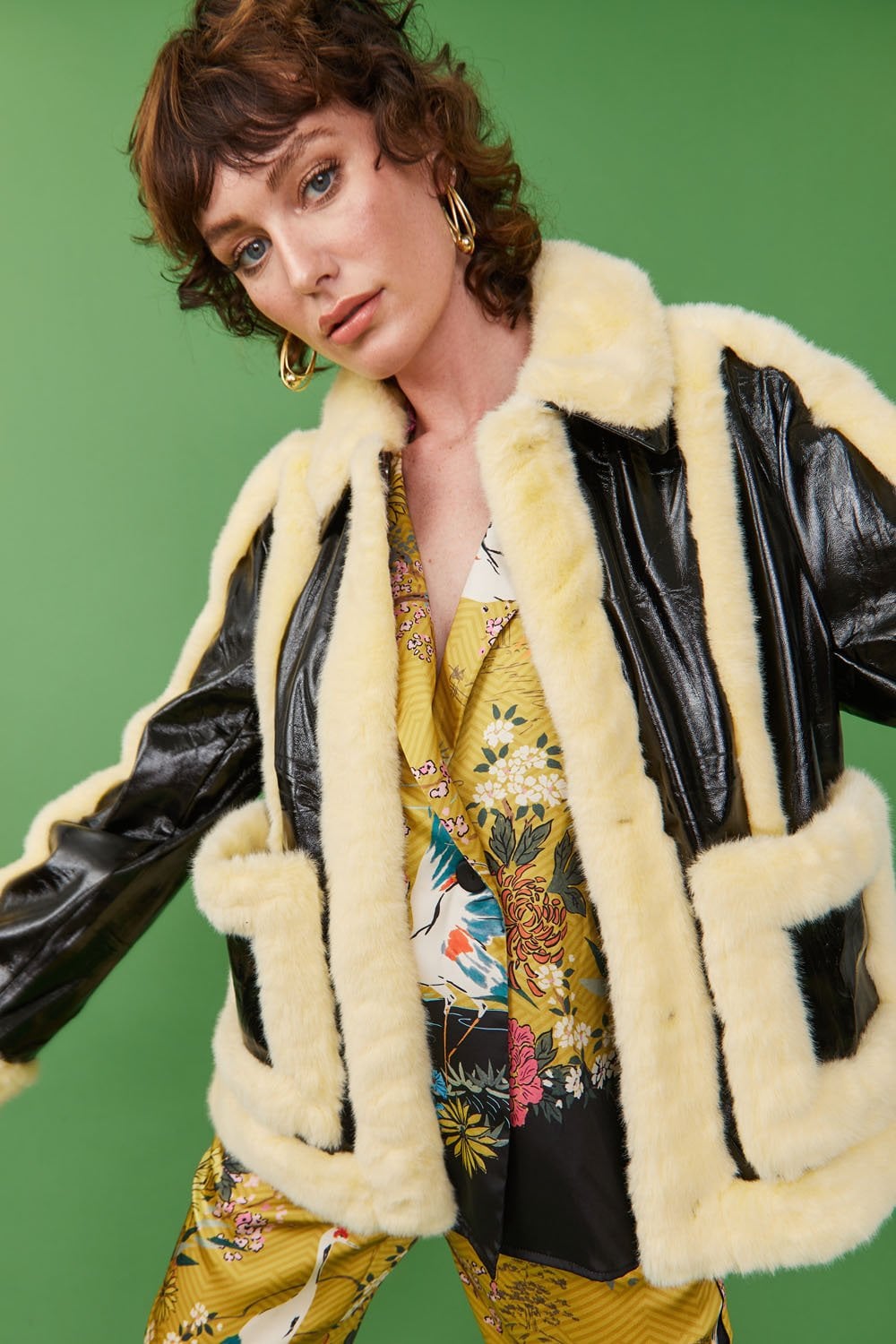 A person poses against a green backdrop, wearing a yellow faux shearling jacket with a shearling collar over a yellow floral outfit.