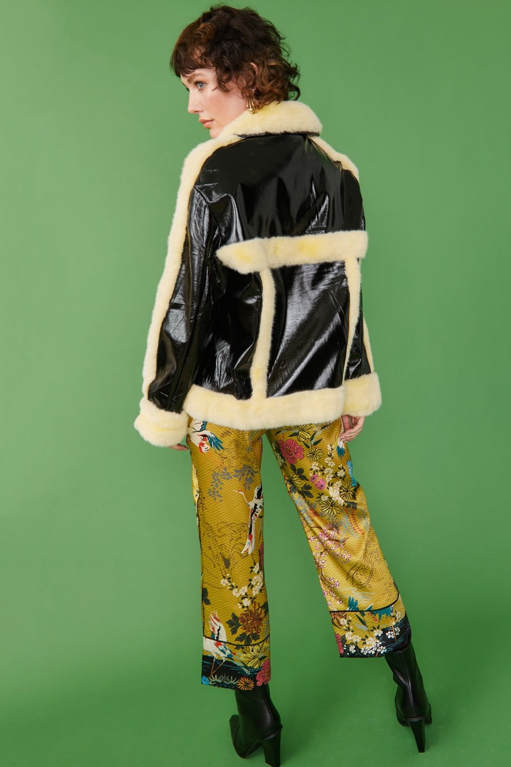 A person stands against a green background wearing a black retro aviator jacket with a white shearling collar, patterned yellow trousers, and black heeled boots.