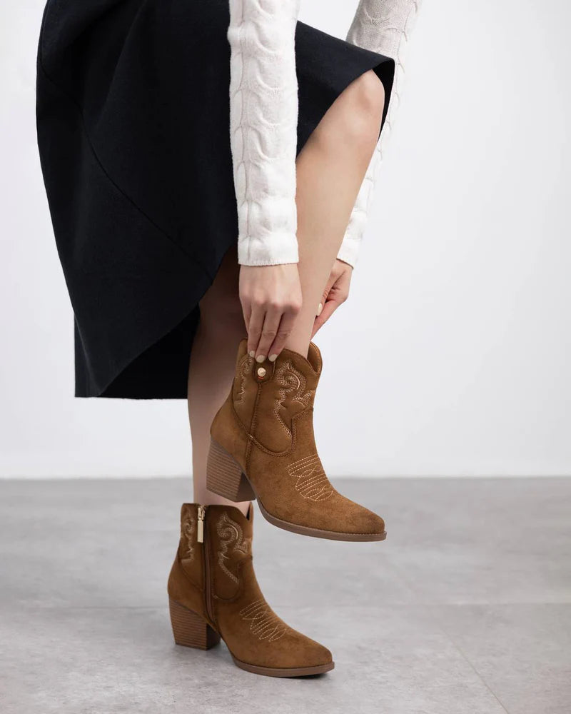 A person in a white textured jumper and black skirt puts on brown suede heeled womens boots featuring embroidered detailing.