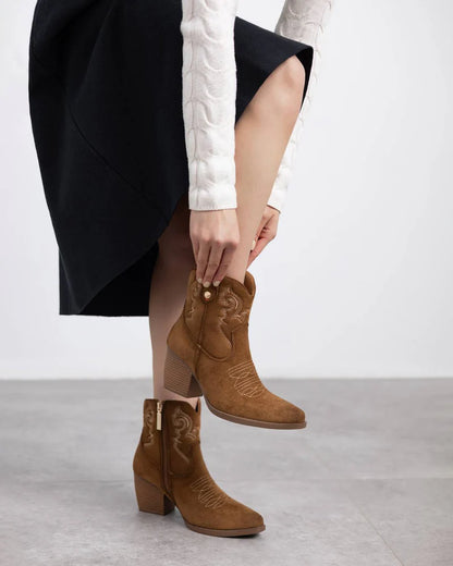 A person in a white textured jumper and black skirt puts on brown suede heeled womens boots featuring embroidered detailing.