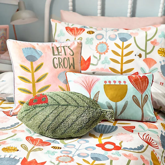 Colourful bed with floral-patterned bedding, a pink cushion featuring LETS GROW text, a rectangular floral cushion, and a nature-inspired cushion shaped like a leaf.