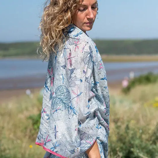 A woman with curly blonde hair wearing a light, patterned kimono-style jacket and a Silver Dragonfly Neck Scarf stands outdoors near water and greenery, glancing over her shoulder.
