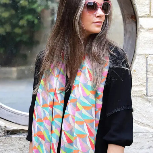 Woman wearing sunglasses and a lightweight summer scarf with vibrant colours and geometric patterns over a black top, standing in front of a round window and stone wall.